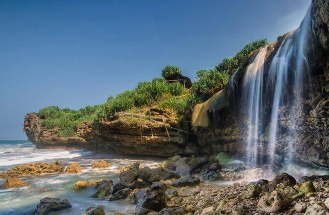 Cascada cayendo sobre un acantilado rocoso hacia el mar, con vegetación verde en la parte alta y cielo despejado